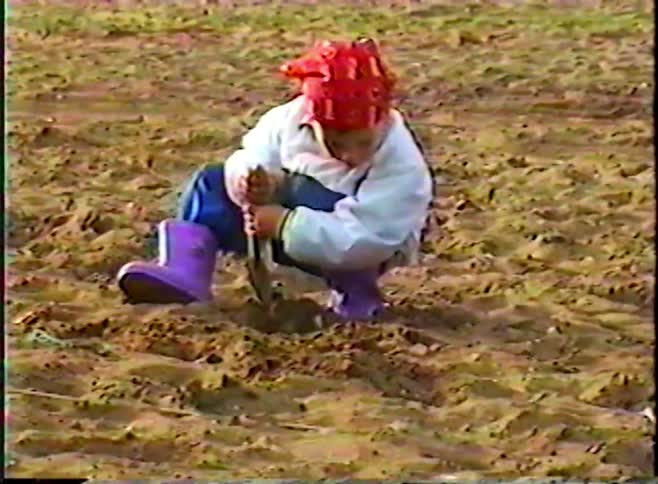 A young child wearing a red bandana and purple rain boots digs in the sand with a trowel on a cool day at the beach