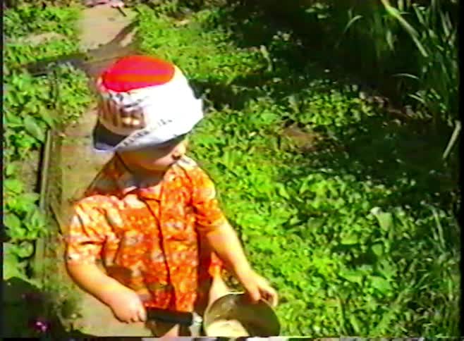 Vintage VHS footage shows a young child in a colorful outfit and hat playing with a pot of water in a lush green backyard garden
