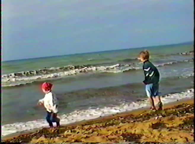 A vintage home video shows two young children playing on the sandy shore as waves crash behind them