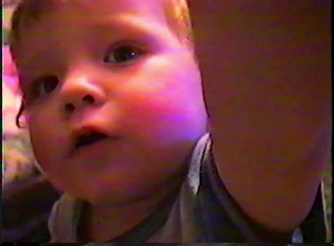 An adorable toddler with blonde hair turns to the camera and opens their mouth in surprise in this vintage home video