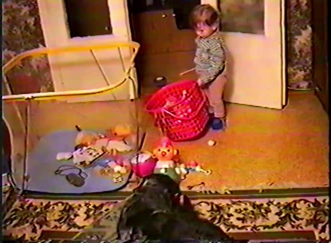 A toddler plays with toys in a messy room as their pet dog eats something off the floor in a vintage home video