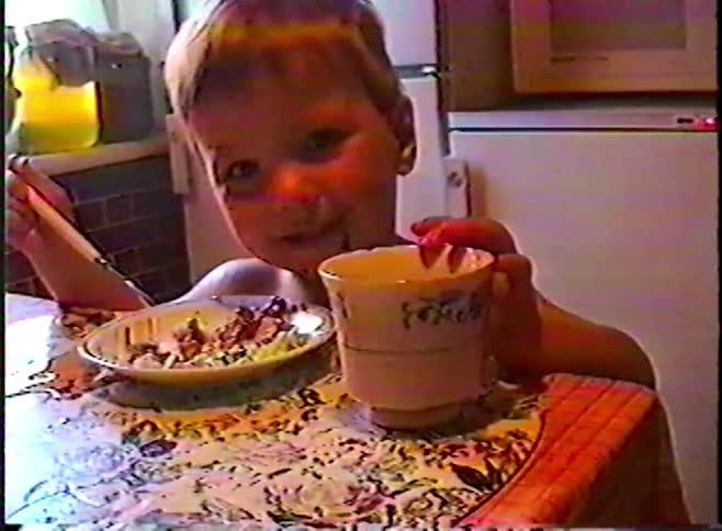 Vintage home video footage shows a cute young boy with a messy face enjoying his meal and a drink at the kitchen table