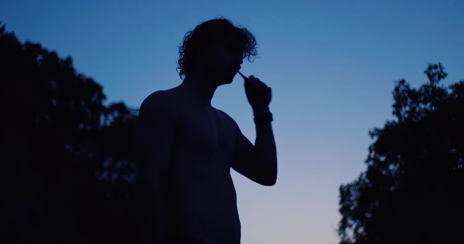 A low-angle silhouette of a shirtless young man with curly hair sucking on a lollipop against a deep blue evening sky