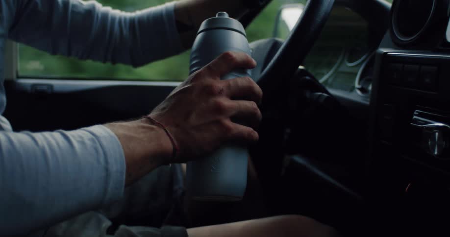 A man holds a reusable water bottle in his hand while driving a car through a scenic, green landscape