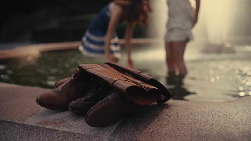 A close-up focuses on discarded shoes and boots as two women spontaneously cool off in a city fountain