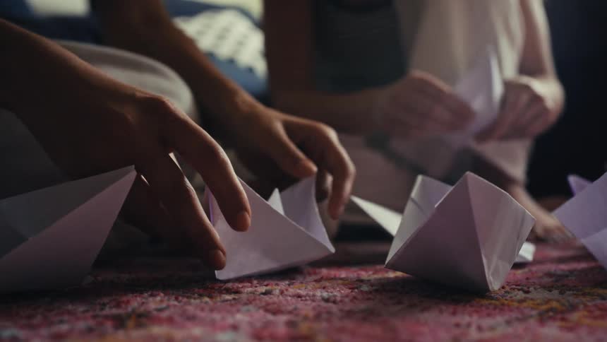 A low-angle close-up captures a fleet of handmade paper boats as two people fold more in the background