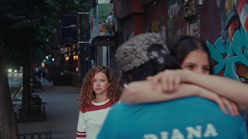 A young woman feels left out as her friend excitedly embraces a man on a graffiti-lined city street at dusk