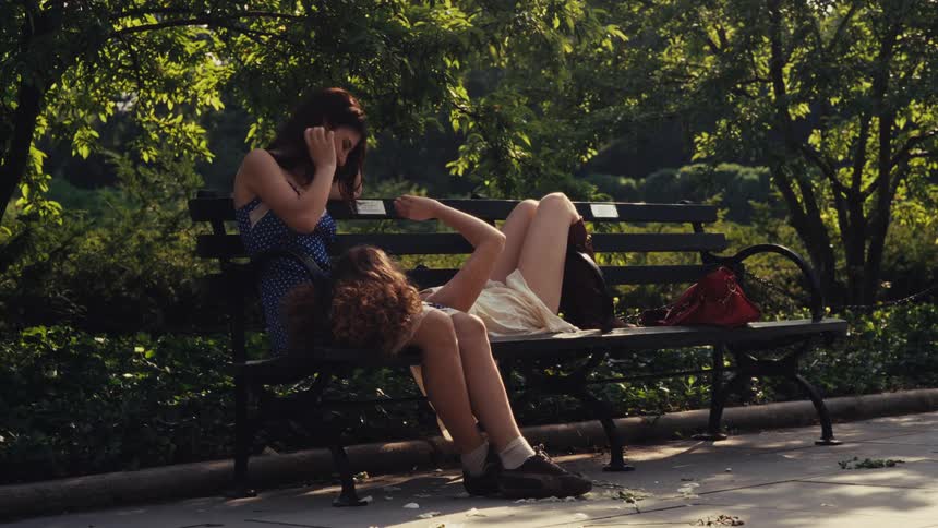 Two young women share an intimate and relaxing moment on a park bench on a sunny day while people walk by
