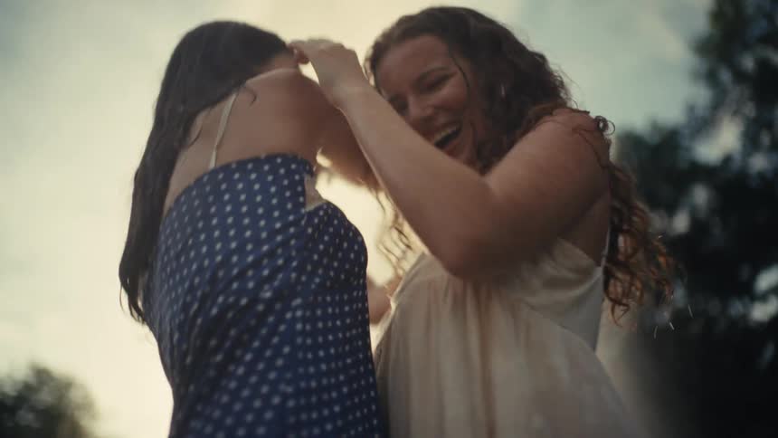 Two carefree young women laugh and dance together in the sunlit mist of a water fountain in slow motion