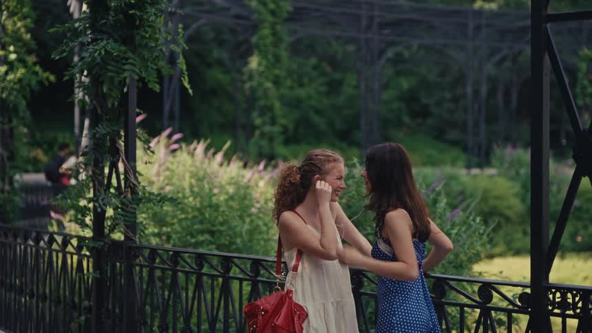 Teenage girls smile and laugh as they share a pair of earbuds to listen to music in a lush city park