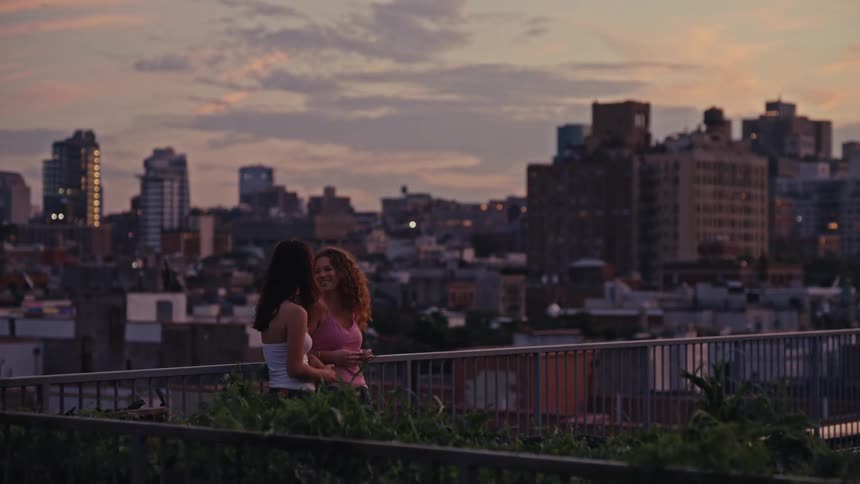A loving lesbian couple shares an intimate kiss and a cigarette on a city rooftop at sunset