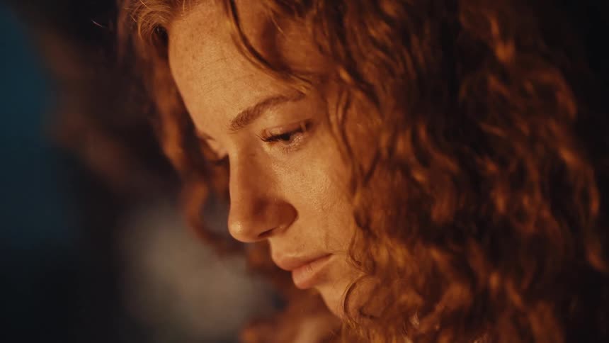 An introspective redhead woman with freckles is lost in thought while writing in a warmly lit room at night