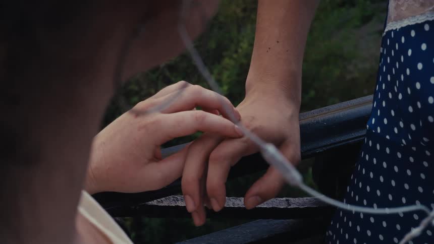 A close-up over-the-shoulder shot of two people's hands tenderly intertwining on a railing as they listen to music together
