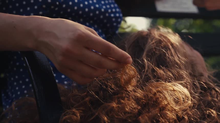 Close up of a mother's hand tenderly playing with her child's curly hair as she rests in her lap on a sunny day