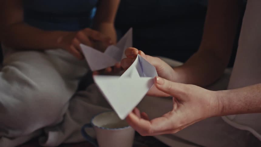 A close-up shot of two people's hands as they sit together and fold white paper into origami boats