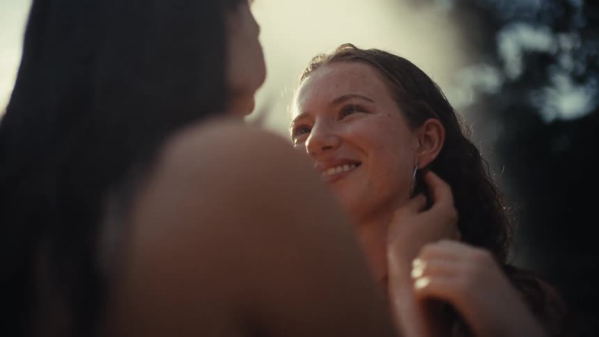 An intimate slow-motion close-up captures a lesbian couple laughing and caressing each other's faces with beautiful sun flares