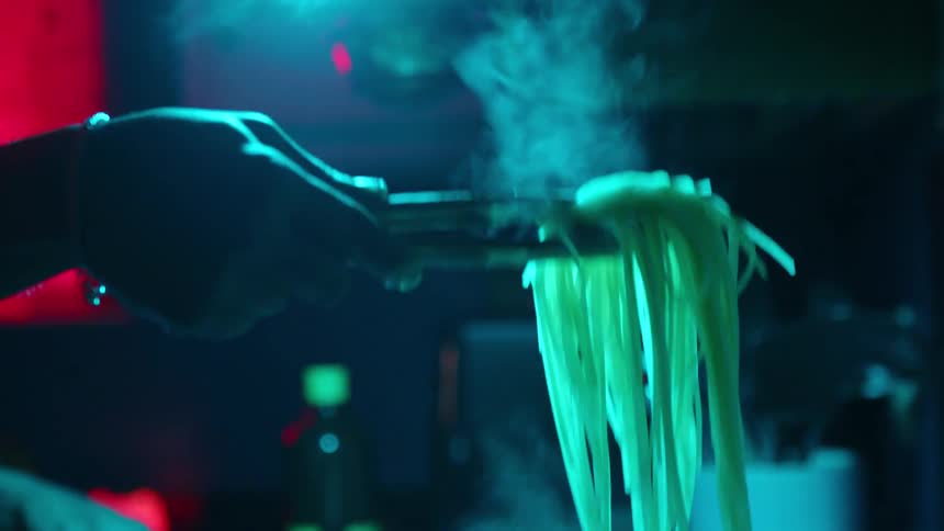 Close up of a tattooed man cooking steaming pasta in a dark kitchen with red and blue lights in slow motion