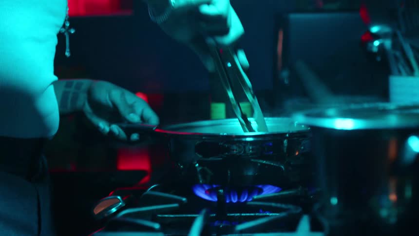 A cook transfers steaming pasta from a pot to a pan on a gas stovetop in a dramatically lit kitchen