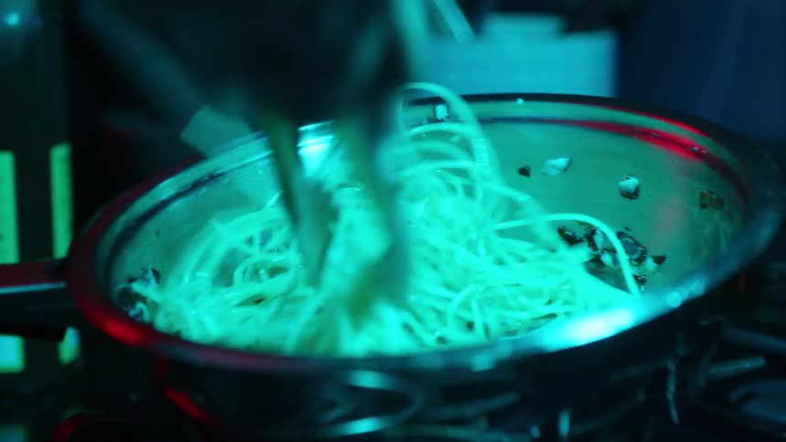 A chef vigorously tosses spaghetti pasta with tongs in a metal pan under a dramatic, moody teal light
