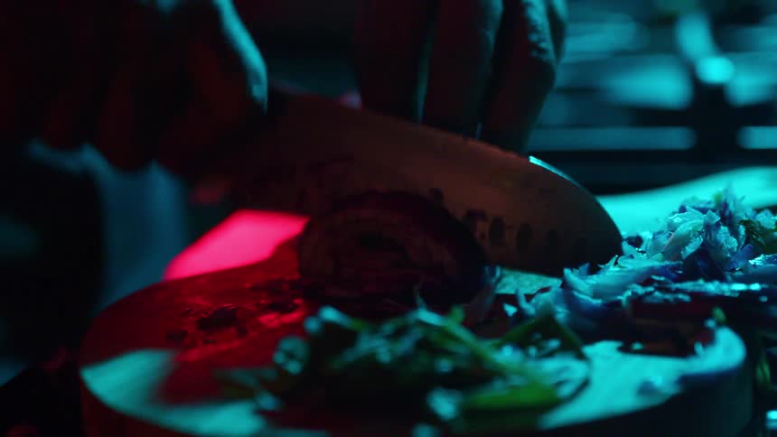 A chef's hands expertly chop a red onion in slow motion on a wooden board under dramatic red and blue lighting