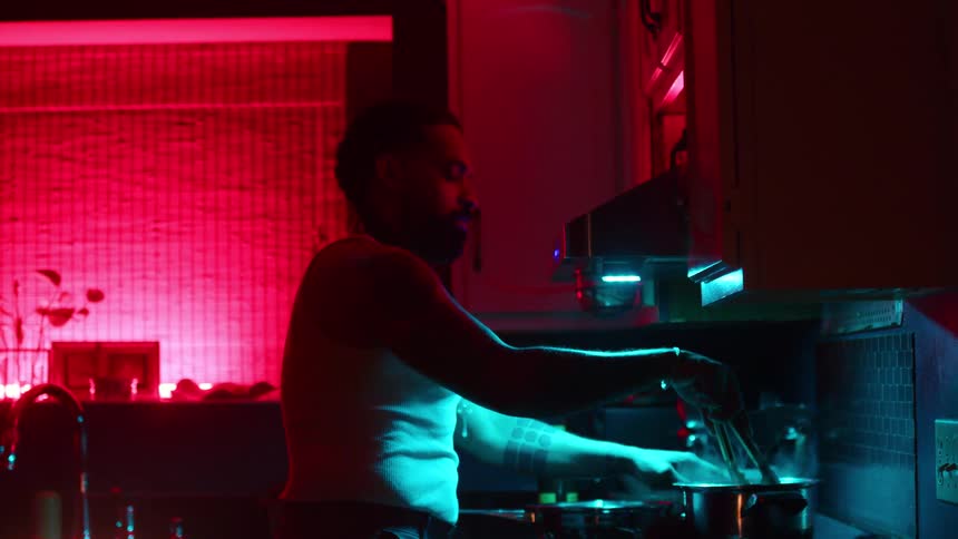 A tattooed man stirs spaghetti in a steaming pot on a gas stove in a stylish kitchen illuminated by red and blue neon lights in slow motion