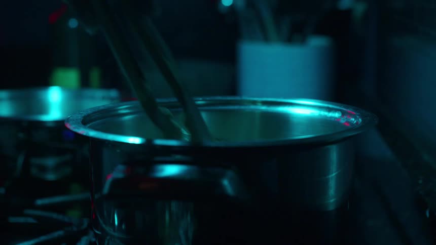 In a dimly lit kitchen, a chef uses tongs to stir pasta as it boils in a steaming pot