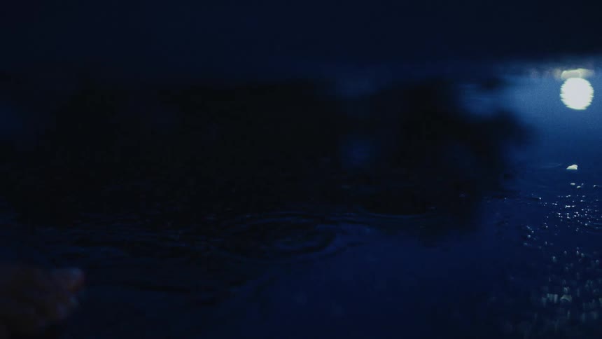 Bare feet splash through a dark puddle at night, disturbing the reflection of a single bright light on the water's surface