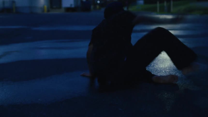 A barefoot man breakdances in a wet, dark parking lot at dusk in slow motion