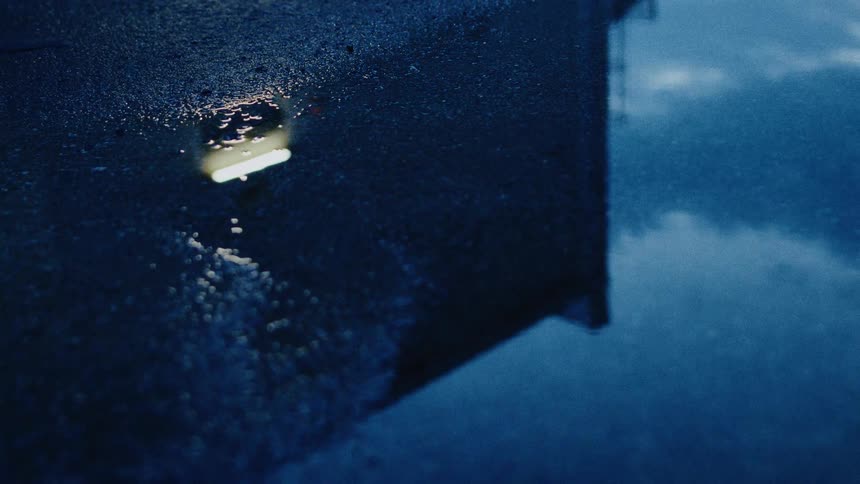 Raindrops fall into a puddle at night, distorting the moody reflection of an illuminated building and the dark blue sky