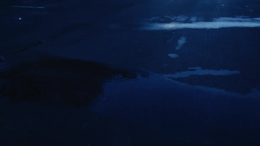 A slow pull-back shot reveals raindrops falling on a dark, wet asphalt parking lot at night
