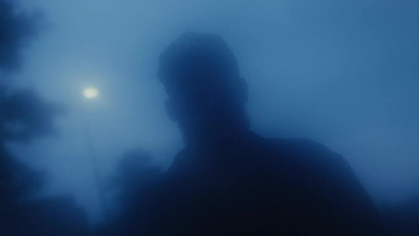 A mysterious figure is silhouetted in the dense blue fog at night, with a single street lamp glowing in the distance