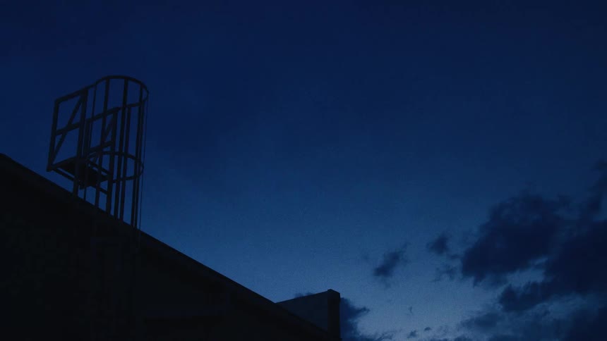 The silhouette of a rooftop access ladder against a moody, deep blue evening sky with soft clouds passing