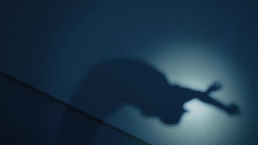 A mysterious and eerie shadow of a person slowly transforms and contorts under a spotlight on a dark wall