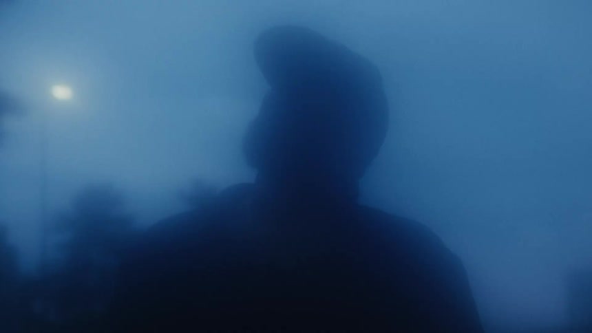 A mysterious, dark silhouette of a man stands in the heavy blue fog under a distant street light