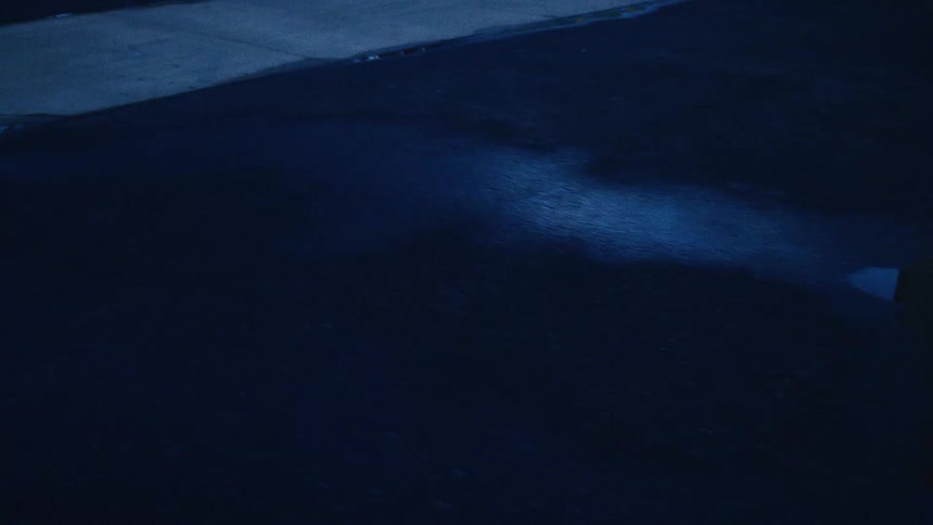 Raindrops fall on a dark, wet surface at night, creating ripples in a puddle as the camera slowly pulls back