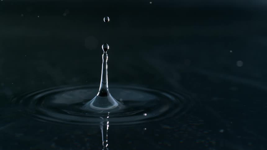 An extreme slow-motion, close-up shot shows a single water droplet falling onto a dark surface, creating a crown splash and radiating ripples