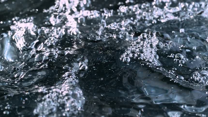 Slow motion closeup shot of dark, bubbling water as more liquid pours in, creating ripples and splashes