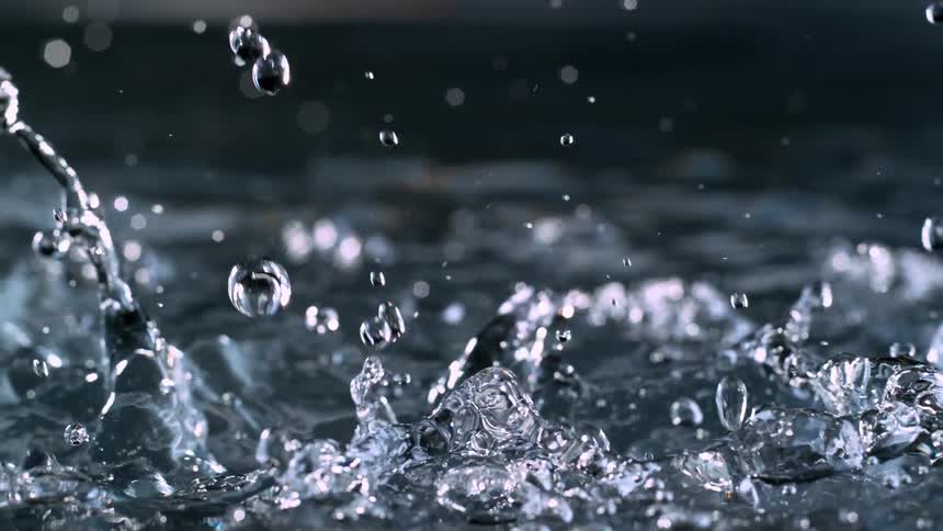 A close-up, slow-motion video captures water droplets falling and splashing into a pool of water, creating beautiful ripples and spray