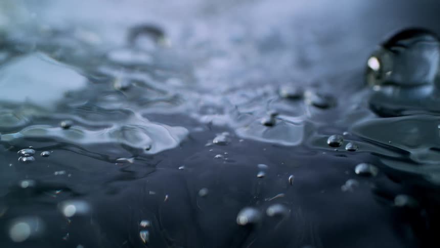 An abstract macro slow-motion shot captures bubbles forming and moving across a dark, undulating water surface