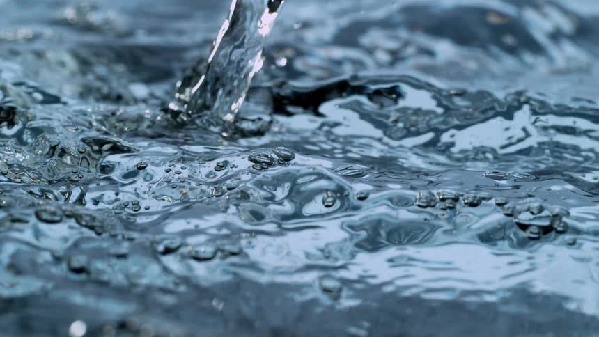 A stream of clear blue water pours into a pool, creating mesmerizing splashes and ripples in a super slow-motion close-up