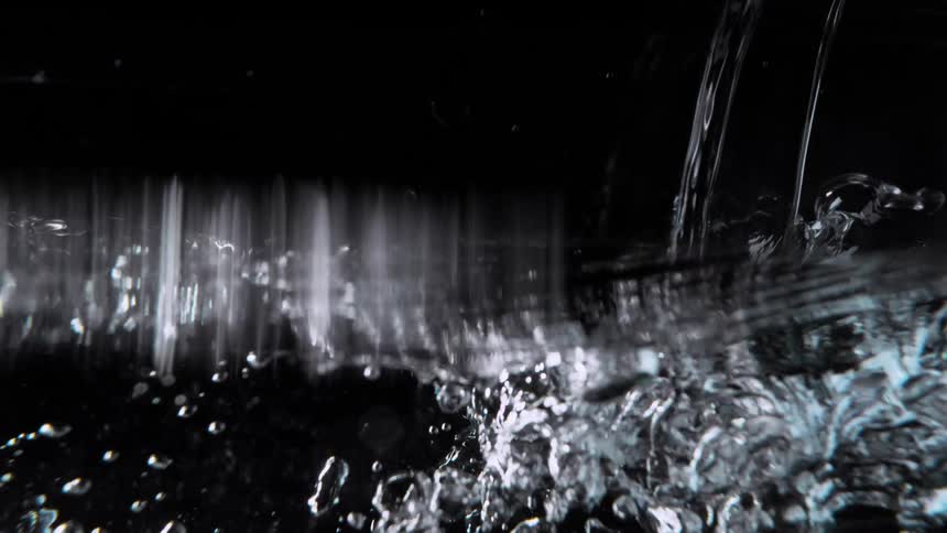 Slow motion close up of clear water being poured, splashing and creating bubbles on a black background until the camera is submerged