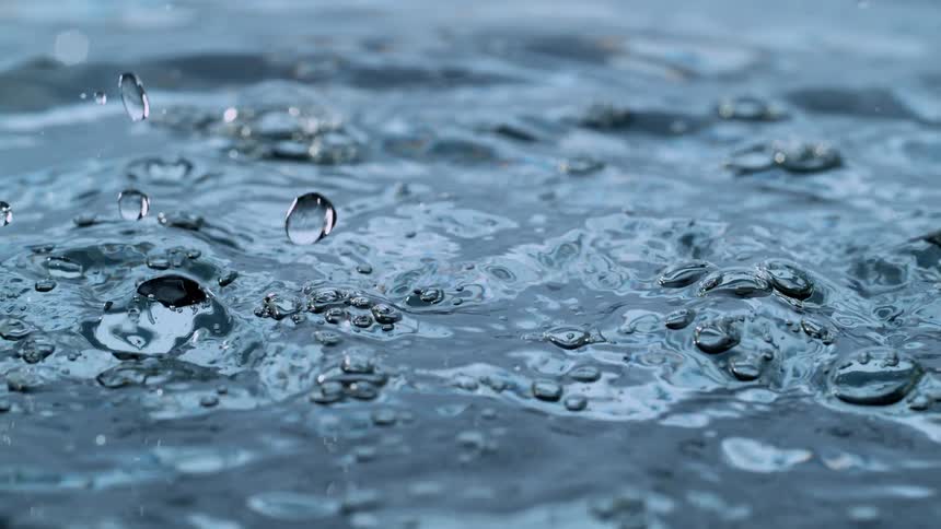 Raindrops fall into rippling water, creating splashes and bubbles in a mesmerizing slow-motion close-up