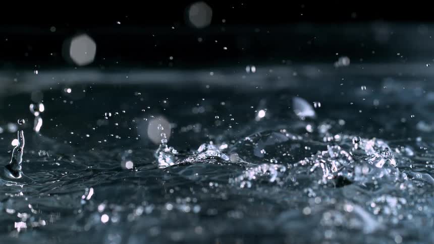 A steady rain of water droplets splashes onto a liquid surface in mesmerizing super slow motion against a black background