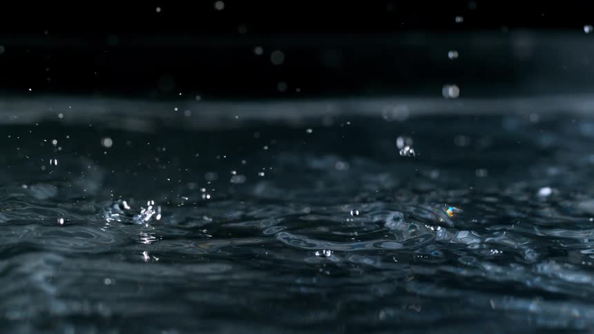 A close-up captures raindrops hitting a water surface in mesmerizing slow motion against a black backdrop