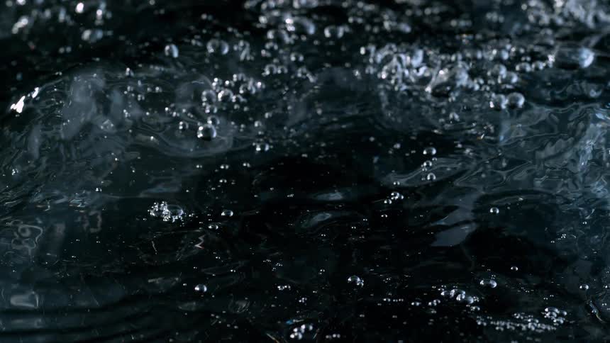 A dramatic close-up captures turbulent water bubbling and forming small crests in extreme slow motion against a black backdrop
