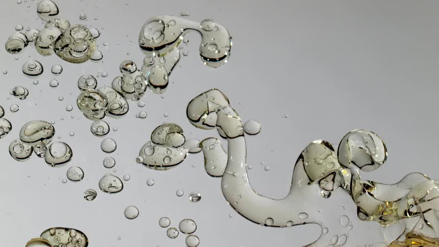A mesmerizing macro shot captures viscous golden oil bubbles slowly combining and separating in a clear liquid against a bright white background