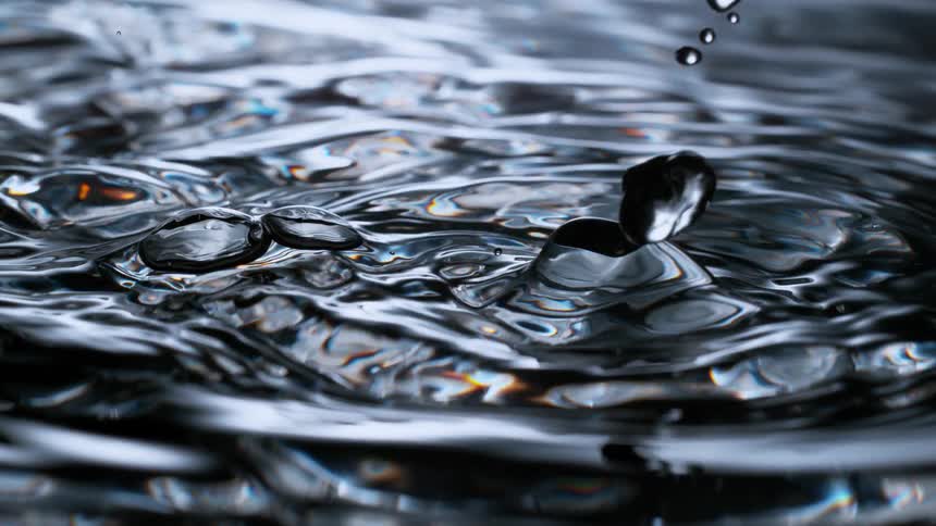A mesmerizing super slow-motion, macro view of a single water droplet making contact with a calm, dark surface, causing a splash and intricate ripples