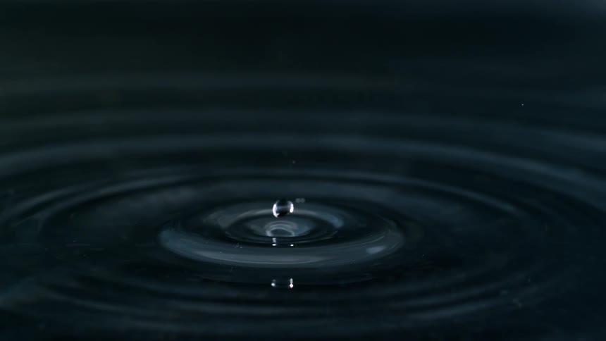 A single water droplet falls in extreme slow motion, creating a mesmerizing splash and perfect ripples on a dark, calm surface