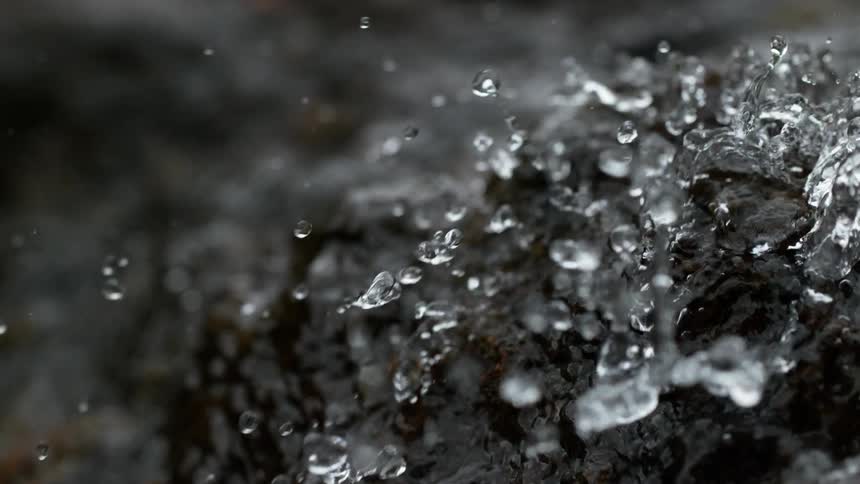 A super slow-motion macro shot captures individual water droplets splashing over a dark, wet rock