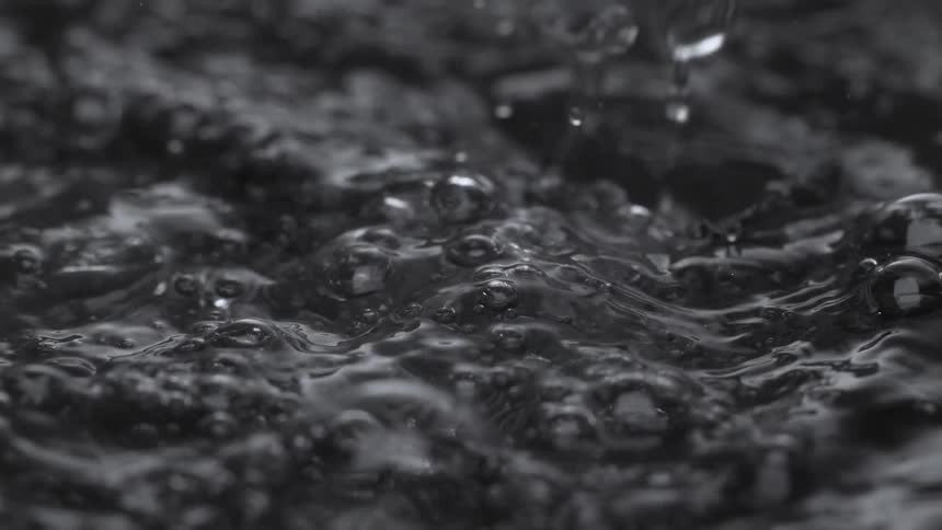 Extreme close up of a stream of dark water pouring into a pool, creating bubbles and ripples in super slow motion